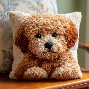 May include: A square cushion with a textured design of a puppy in shades of brown and cream. The puppy has dark eyes and a black nose. The cushion is placed on a wooden surface.