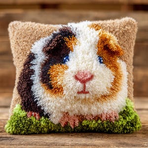 Op de afbeelding: Een vierkant, beige sierkussen met een gedetailleerd, getextureerd ontwerp van een cavia. De cavia heeft bruine, witte en oranje vacht, blauwe ogen en een roze neus. Groene plukjes garen vormen het gras aan de onderkant van het kussen.