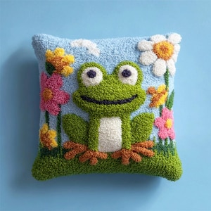 May include: A square throw pillow featuring a textured punch needle design of a green frog with a white belly, centered against a light blue background. The scene includes colorful yellow, pink, and white flowers and small white clouds.