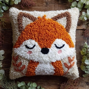 May include: A square throw pillow featuring a textured punch needle design of a sleeping fox face. The fox has orange fur, white cheeks, a black nose, closed eyes, and pink blush. The background is cream with orange leaf accents.