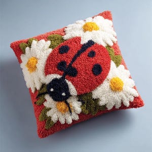 以下が含まれることがあります： 赤いテントウムシと白いデイジーの花がパンチニードル技法で刺繍された正方形のクッション。テントウムシは黒い斑点と頭部を持ち、デイジーは黄色い中心部と緑の葉が特徴で、全体がループ状の糸で構成された赤い背景のテキスタイルです。