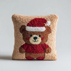 Peut inclure: Un coussin carré à la surface texturée et touffetée représentant un ours en peluche marron portant un pull rouge et un bonnet de Père Noël rouge avec un pompon blanc. L'ours a un museau crème, des yeux sombres et un petit nez sur fond beige.