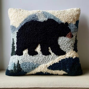 Peut inclure: Un coussin carré avec un motif en punch needle texturé représentant un grand ours noir centré sur un paysage de montagnes et de sapins dans des tons crème, bleu clair et bleu marine. L'ours présente un petit détail marron au museau.