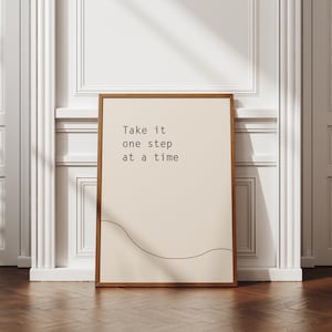 Könnte beinhalten: Gerahmter Druck mit dem Text "Take it one step at a time" in minimalistischem Stil. Der Druck hat einen hellbeigen Hintergrund mit einer dünnen, schwarzen, geschwungenen Linie am unteren Rand. Der Rahmen ist warmbraun.