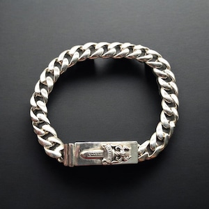 May include: A silver curb chain bracelet with a rectangular clasp. The clasp features a dagger and ornate floral design. The bracelet is made of thick, interlocking links and is displayed on a black background.