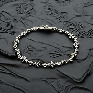 May include: Silver bracelet with a daisy flower design. The bracelet is made up of daisy-shaped links connected by small, circular links. The clasp is a simple hook-and-eye style. The bracelet is displayed on a textured black surface.