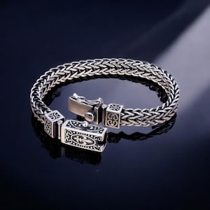 May include: A silver-colored bracelet with a woven chain design. The bracelet features decorative square clasps with ornate patterns and a secure clasp closure. The jewelry is displayed on a dark surface.