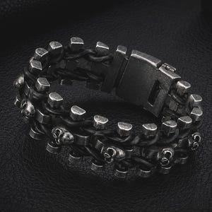 以下が含まれることがあります： スカルデザインのダークグレーのブレスレット。ブレスレットは、編み込まれた黒い革のストランドでできており、小さなシルバー色のスカルと長方形の金属アクセントが特徴です。留め金は、お揃いのダークグレーの金属です。