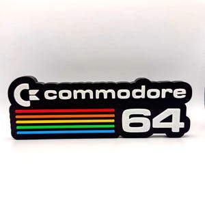 Può includere: Un logo nero Commodore 64 con testo bianco e un design a strisce arcobaleno. Il logo presenta il testo "Commodore 64" in un carattere stilizzato. Lo sfondo è bianco.