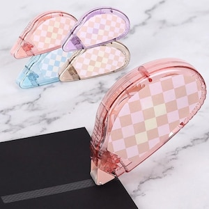 May include: A collection of pastel-colored correction tape dispensers. The dispensers are teardrop-shaped with a checkered pattern and are in shades of pink, purple, blue, and beige. One dispenser is shown in use, correcting a black surface.