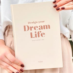 Könnte beinhalten: Ein cremefarbenes Notizbuch mit dem Text "Design your Dream Life" in einem rötlich-braunen Farbton. Das Notizbuch enthält auch die Wörter "dream - envision - plan" und das Wort "FLOW". Das Notizbuch wird von einer Person gehalten.
