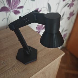 May include: A black desk lamp with an adjustable arm and a hexagonal base. The lamp has a cylindrical head and a conical shade. The lamp is made of a matte black material and sits on a wooden surface.
