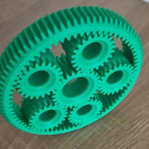 Può includere: Un set di ingranaggi planetari verde brillante stampato in 3D. Il set comprende una grande corona dentata esterna con denti interni e diversi ingranaggi più piccoli. Gli ingranaggi sono progettati per ingranare per la funzione meccanica.