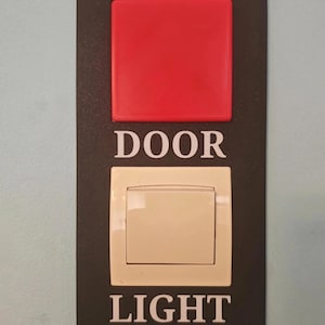 Peut inclure: Panneau rectangulaire noir avec un bouton carré rouge en haut et un interrupteur de lumière crème en bas. Les mots "DOOR" et "LIGHT" sont imprimés en blanc, respectivement au-dessus et en dessous de l'interrupteur.