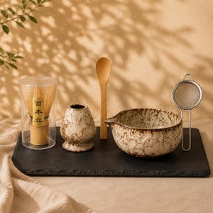 May include: A matcha tea set including a bamboo whisk in a clear cylindrical case with Japanese text, a speckled ceramic whisk holder, a wooden spoon, a speckled ceramic pouring bowl, and a small metal mesh strainer on a black slate board.