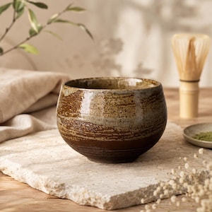 Peut inclure: Un bol à matcha en céramique fait à la main, avec un glaçage texturé brun et beige. Le bol est rond avec une large ouverture. Un fouet en bambou et une petite coupelle de poudre de matcha suggèrent une cérémonie du thé.