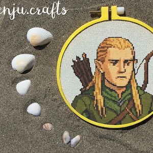 May include: A cross-stitch portrait of a character with blonde hair, wearing a green tunic, and carrying a bow and arrows, set in a yellow hoop. The image is surrounded by seashells on a sandy surface.