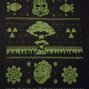 May include: A black fabric piece featuring a green cross-stitch pattern. The design includes a power armor helmet, two bombs, two radiation symbols, a mushroom cloud, a smiling character face, and two gear icons arranged in horizontal rows.