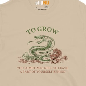 May include: Tan t-shirt with a green snake graphic and the text "TO GROW, YOU SOMETIMES NEED TO LEAVE A PART OF YOURSELF BEHIND." The snake is coiled and has a detailed design. The shirt has a crew neck and short sleeves.