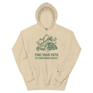 Op de afbeelding: Een beige hoodie met een groene afbeelding van een wolf, beer en eland met een berg op de achtergrond. De tekst "FIND YOUR PATH TO YOUR INNER BEAST" staat eronder. De hoodie heeft een voorzak en een capuchon.