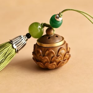 Può includere: Un piccolo bruciatore di incenso in legno con un design a fiore di loto intagliato. Il bruciatore ha un bordo color ottone e una piccola perla di giada verde attaccata a un cordino verde. Un nappa verde pende dal lato, aggiungendo un elemento decorativo.