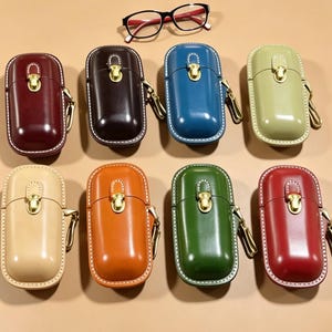 Può includere: Una collezione di otto custodie in pelle di vari colori, tra cui bordeaux, marrone, blu e verde. Ogni custodia ha una chiusura dorata e un moschettone abbinato. Un paio di occhiali con montatura nera e dettagli rossi è sopra.