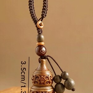 May include: A brown wooden gourd-shaped pendant featuring intricate carved swirl patterns. It includes a braided cord loop, two light-colored wooden beads, and a cluster of four dark grey beads. The pendant measures 1.37 inches in height.