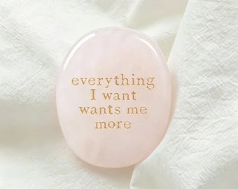 May include: A smooth, oval-shaped pale pink rose quartz stone featuring the gold-colored engraved text "everything I want wants me more" centered on the front surface. The stone has a polished, glossy finish and a translucent appearance.