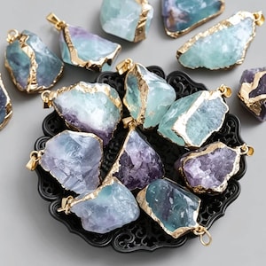 Può includere: Una collezione di pendenti in fluorite grezza con bordi color oro. I pendenti presentano una miscela di tonalità blu, verdi e viola. Sono disposti su un piatto decorativo nero, mettendo in risalto la loro bellezza naturale e le forme uniche.