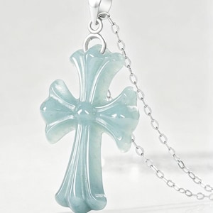 Può includere: Collana con pendente a forma di croce azzurra. Il pendente ha una forma a croce stilizzata con bordi arrotondati e superficie lucida. È appeso a una delicata catena d'argento. Lo sfondo è bianco neutro.