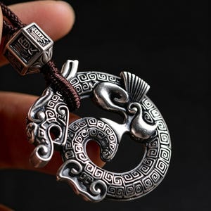 Può includere: Un ciondolo color argento con un design a drago avvolto, caratterizzato da intricati motivi geometrici e una finitura liscia e lucida. Il ciondolo è sospeso a un cordino intrecciato marrone scuro, che passa attraverso una perlina quadrata color argento con dettagli incisi.
