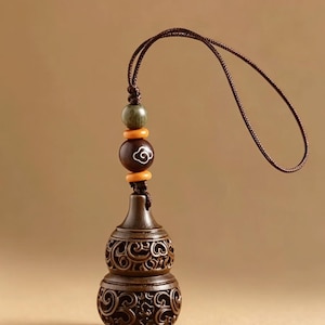 May include: A dark brown wooden gourd-shaped pendant featuring intricate openwork scroll carvings. The pendant is attached to a brown cord with a dark green bead, two small orange spacer beads, and a larger brown bead with a white cloud motif.