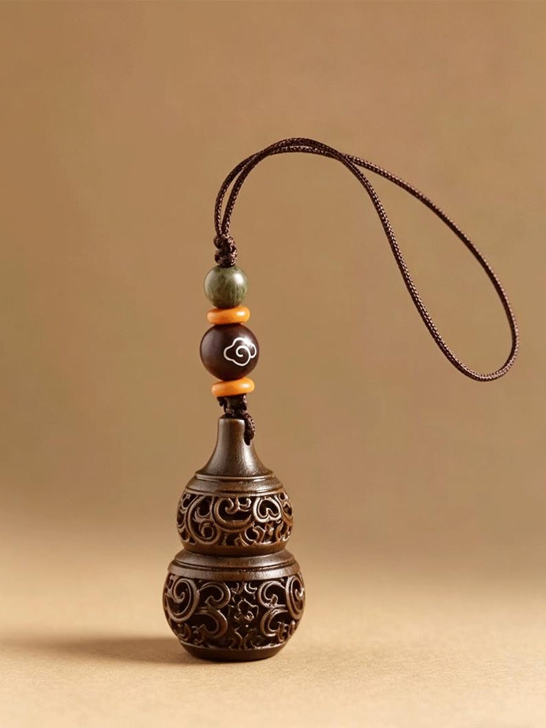 May include: A dark brown wooden gourd-shaped pendant featuring intricate openwork scroll carvings. The pendant is attached to a brown cord with a dark green bead, two small orange spacer beads, and a larger brown bead with a white cloud motif.