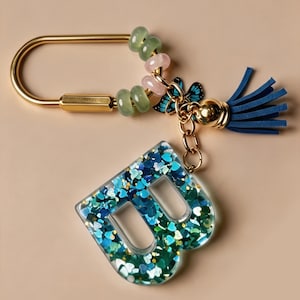 Può includere: Un portachiavi in metallo dorato con un ciondolo a forma di lettera B riempito di glitter blu e verdi a forma di cuore. Il portachiavi include un moschettone dorato, perline verde chiaro e rosa, un piccolo ciondolo a farfalla blu e una nappa in pelle blu.