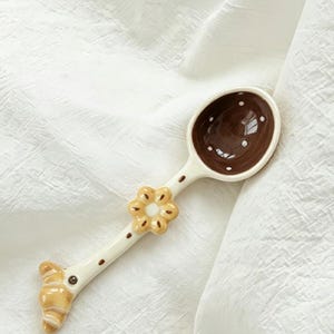 Pode incluir: Uma colher decorativa de cerâmica com uma tigela castanha e bolinhas brancas. O cabo é branco com bolinhas castanhas, um detalhe floral e uma extremidade em forma de croissant. A colher está sobre uma superfície branca texturizada.