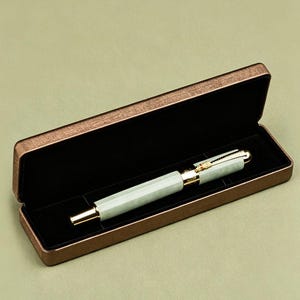 May include: A luxury fountain pen with a light green barrel and gold-colored accents rests inside a brown pen case with a black interior. The pen case is open, revealing the pen's design. The background is a solid olive green.