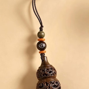 May include: A dark brown wooden gourd-shaped pendant featuring intricate openwork scroll carvings. The ornament hangs from a brown cord adorned with a green wooden bead, two small orange spacer beads, and a dark bead with a white cloud motif.