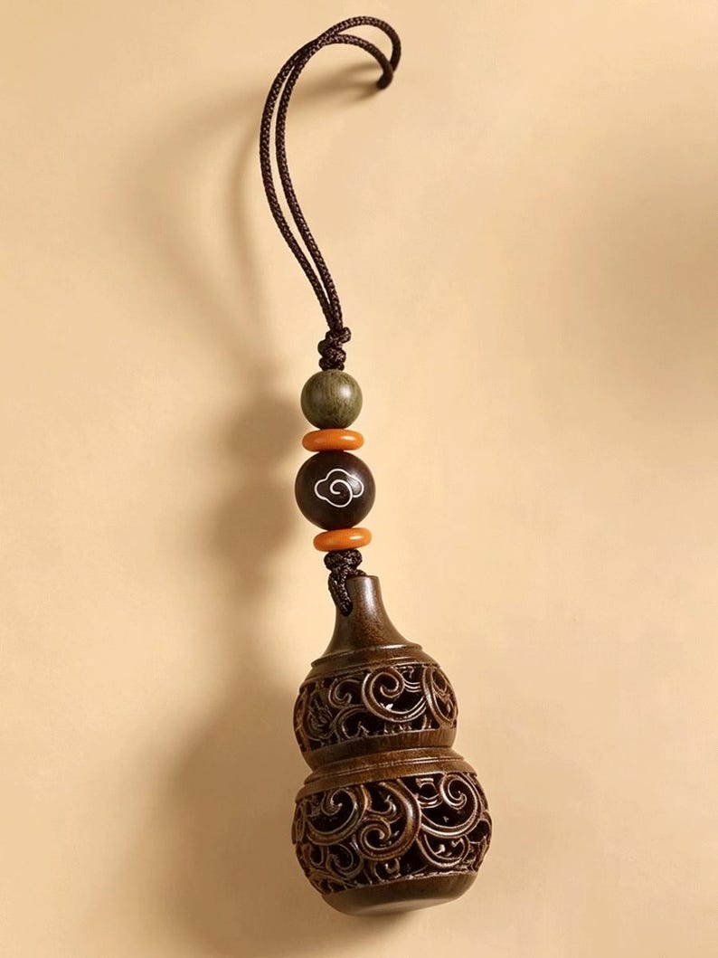 May include: A dark brown wooden gourd-shaped pendant featuring intricate openwork scroll carvings. The ornament hangs from a brown cord adorned with a green wooden bead, two small orange spacer beads, and a dark bead with a white cloud motif.