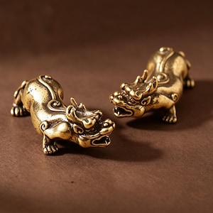 Può includere: Due piccole statuette di drago in ottone. I draghi sono in posizione accovacciata con la bocca aperta, mostrando squame e dettagli intricati. Lo sfondo è marrone caldo, che mette in risalto il colore dorato delle statuette.