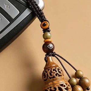 May include: A brown wooden gourd-shaped charm with intricate openwork carvings, attached to a dark brown braided cord. The cord features several small wooden beads in shades of tan, olive green, and dark brown, with one bead displaying a white symbol.