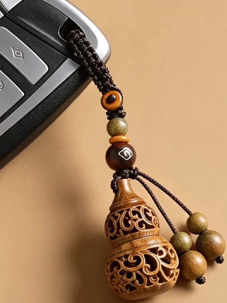May include: A brown wooden gourd-shaped charm with intricate openwork carvings, attached to a dark brown braided cord. The cord features several small wooden beads in shades of tan, olive green, and dark brown, with one bead displaying a white symbol.