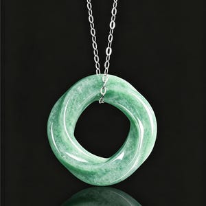 May include: A green jade pendant necklace with a silver chain. The pendant is a smooth, rounded square shape with a hole in the center. The jade has a translucent quality and a subtle variation in color. The necklace hangs against a black background.