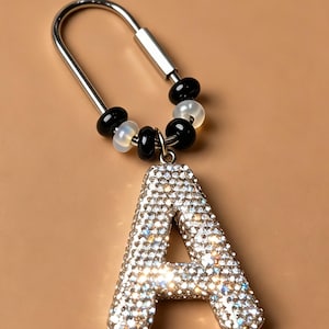 Può includere: Un portachiavi a forma di U color argento con un ciondolo a forma di lettera A ricoperto di strass trasparenti scintillanti. L'anello metallico è decorato con perline alternate nere e bianche traslucide, creando un accessorio a contrasto.