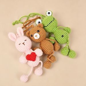 May include: Three crocheted plush toys including a light pink rabbit with a red heart, a brown bear, and a green frog. Each toy features embroidered facial details, dangling limbs, and a long green cord attached to the top for hanging purposes.