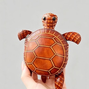 Può includere: Un portamonete a forma di tartaruga in pelle marrone con chiusura a cerniera. La tartaruga ha un motivo a guscio strutturato e dettagliato e piccoli occhi rotondi. La borsa è tenuta su uno sfondo grigio chiaro.