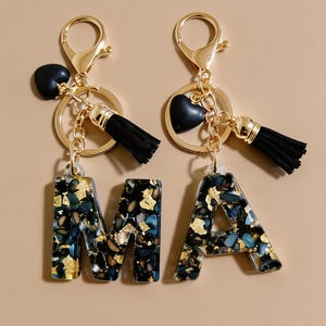 Può includere: Due portachiavi a forma di lettere M e A, realizzati in resina scura con scaglie blu, nere e dorate incorporate. Ogni portachiavi include un moschettone in metallo dorato, un anello, un ciondolo a forma di cuore nero e una nappa nera.