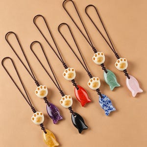 Op de afbeelding: Een collectie van zeven telefoonhangers, elk voorzien van een crèmekleurige kattenpootkraal gecombineerd met een gladde, visvormige hanger in diverse kleuren zoals geel, paars, zwart, rood, blauw, groen en lichtroze, bevestigd aan bruine koorden.