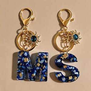 Può includere: Due portachiavi a forma di lettere M e S, realizzati in resina blu con frammenti di pietra incastonati e scaglie di foglia d'oro. Ogni portachiavi include un moschettone in metallo dorato, un anello circolare e un ciondolo a forma di sole.