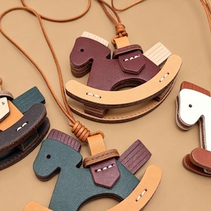 Può includere: Collane con ciondoli a forma di cavallo a dondolo in pelle in vari colori: bordeaux, verde e bianco. Ogni ciondolo presenta un design a cavallo a dondolo con una sella ed è attaccato a un cordino di cuoio marrone. Le collane sono su uno sfondo neutro.