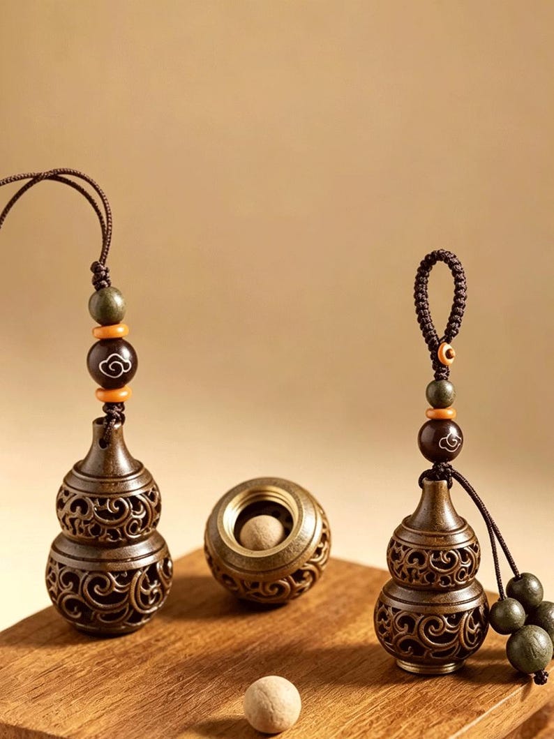 May include: Two bronze-colored metal gourd-shaped containers with intricate openwork scroll patterns, each featuring a brown cord with dark green and orange beads. A small spherical container with a matching scroll design and a light-colored ball are present.